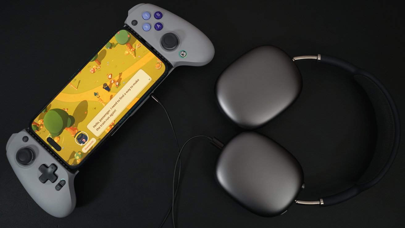 iPhone в контроллере GameSir показывает сцену из «Тамагатчи», красочной игры, в которой желтый персонаж идет по лесу. Рядом с контроллером лежит пара серых AirPods Max.