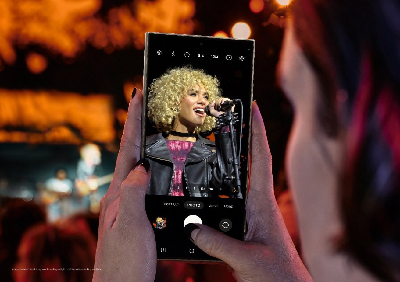 Руки держат смартфон, на котором запечатлено выступление певца на сцене, видно через экран камеры телефона, на фоне размытой публики.