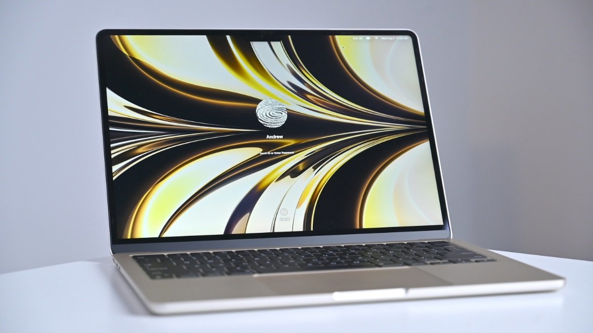 MacBook Air на белом столе с яркими абстрактными обоями с завитками, приглашением для входа в систему с надписью «Эндрю» и значком отпечатка пальца.