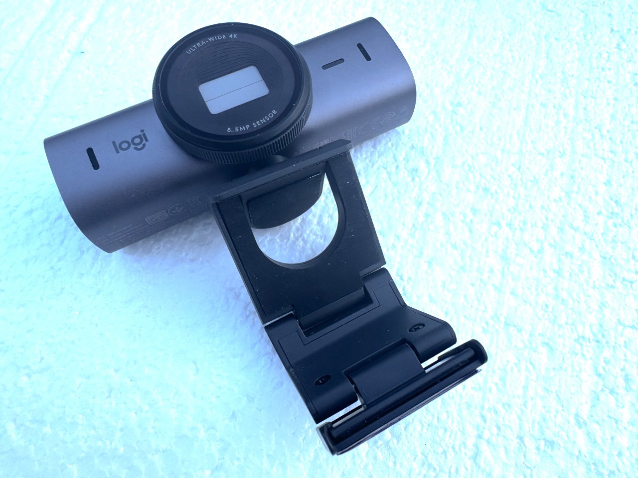Веб-камера Logitech 4K с крышкой объектива, закрепленная на черном зажиме для монитора на текстурированном голубом фоне.