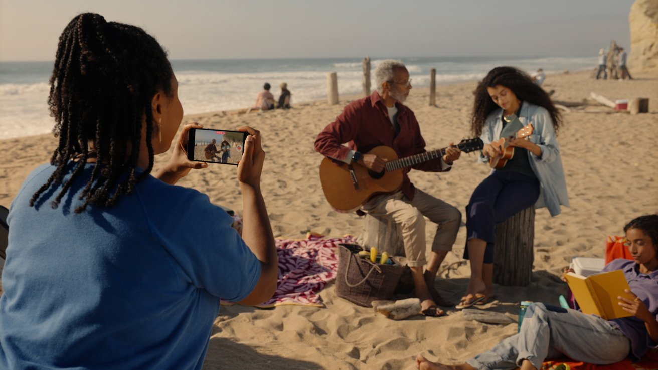 Женщина записывает на свой iPhone видео, на котором два человека играют на музыкальных инструментах на пляже.