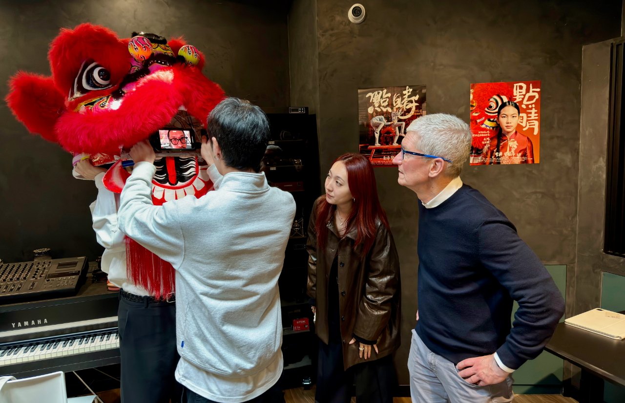 Три человека в помещении в красном танцевальном костюме льва, один держит телефон, на котором изображено мужское лицо, рядом музыкальные инструменты и культурные плакаты на стене.