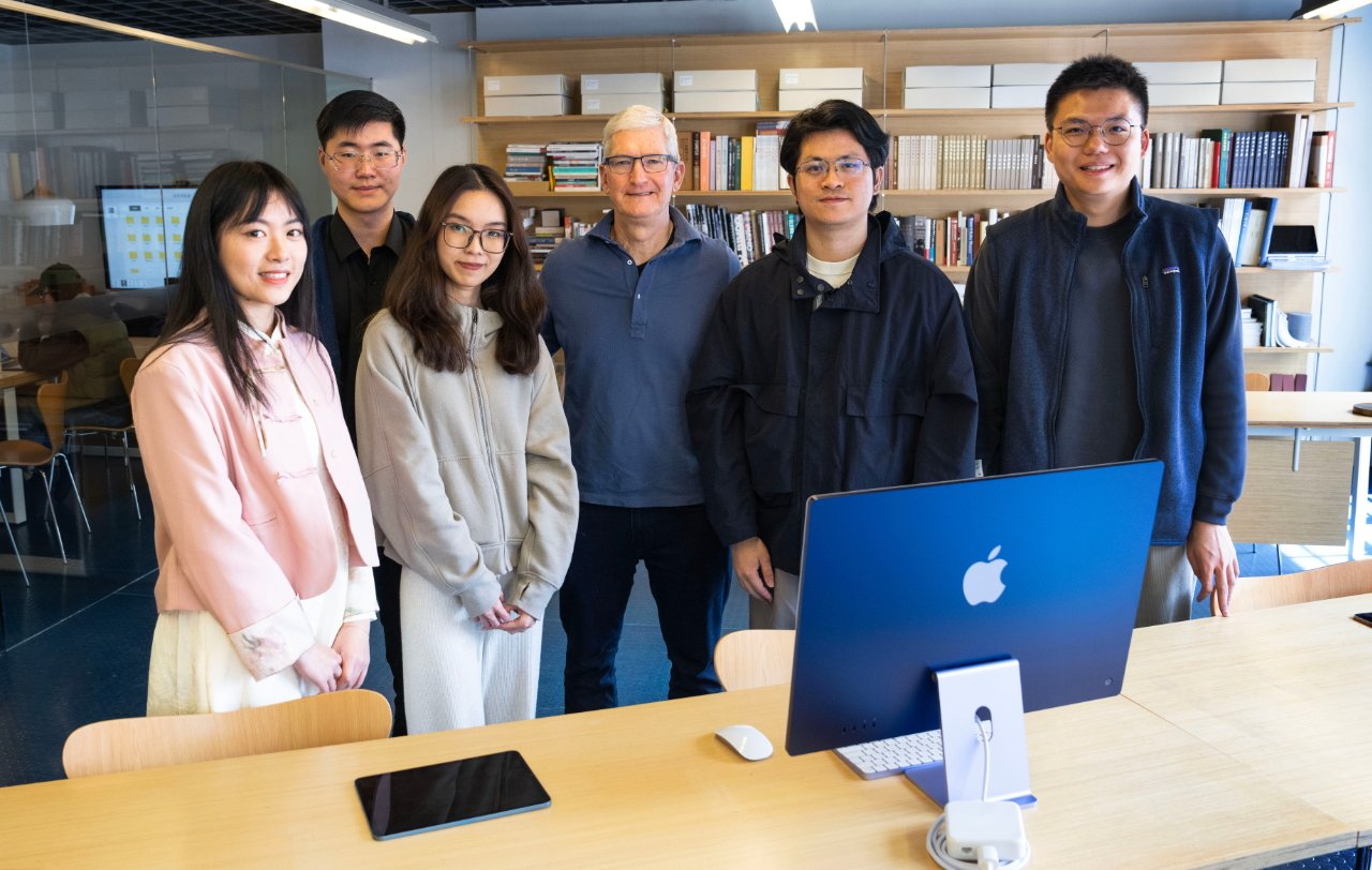Группа из пяти человек улыбается за столом с компьютером Apple в современном офисе.
