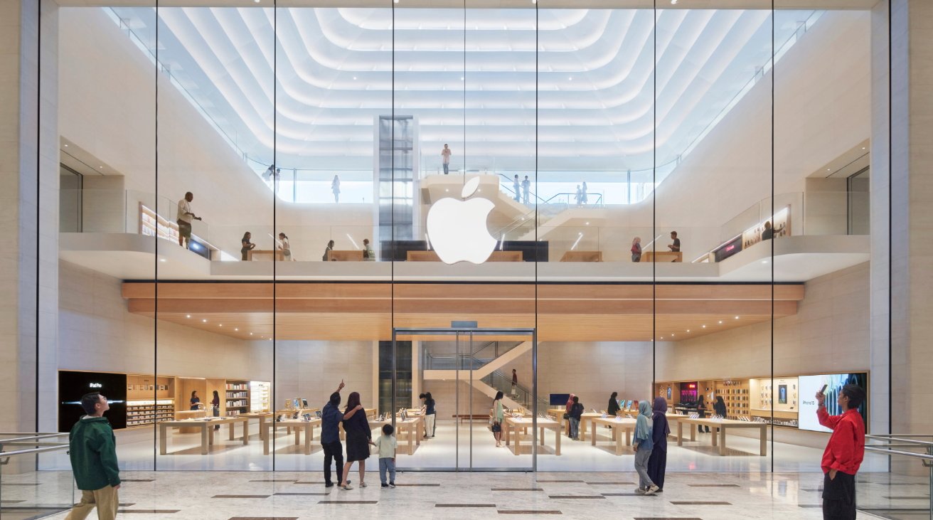 Современный двухэтажный магазин с большим стеклянным фасадом, подсвеченным логотипом яблока, разнообразными витринами с товарами и несколькими покупателями, просматривающими внутри.