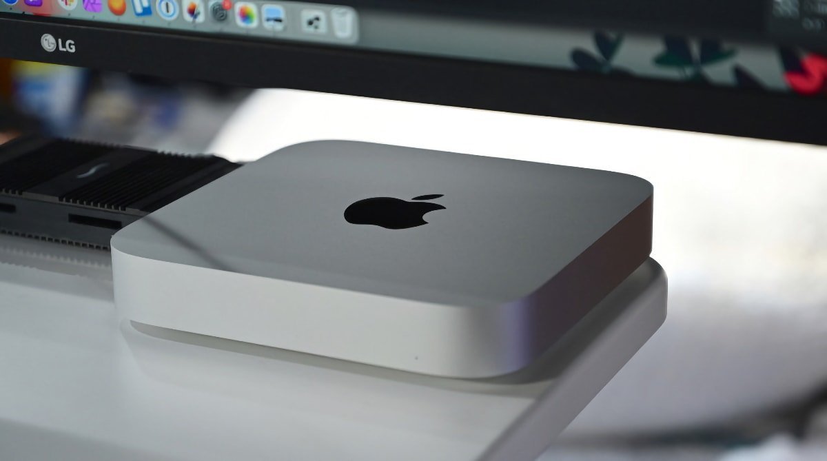 Серебристый компьютер Apple Mac Mini на столе, на заднем плане — монитор LG, на котором отображается рабочий стол macOS.