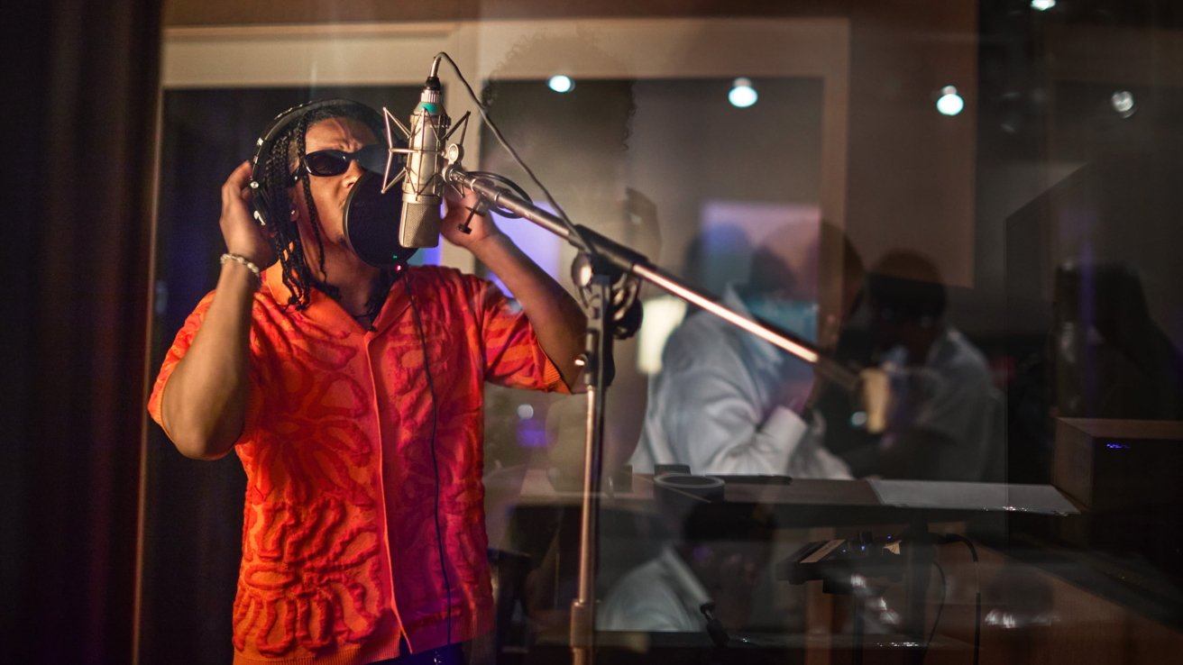 Человек в красной рубашке и солнечных очках поет в микрофон в студии звукозаписи.